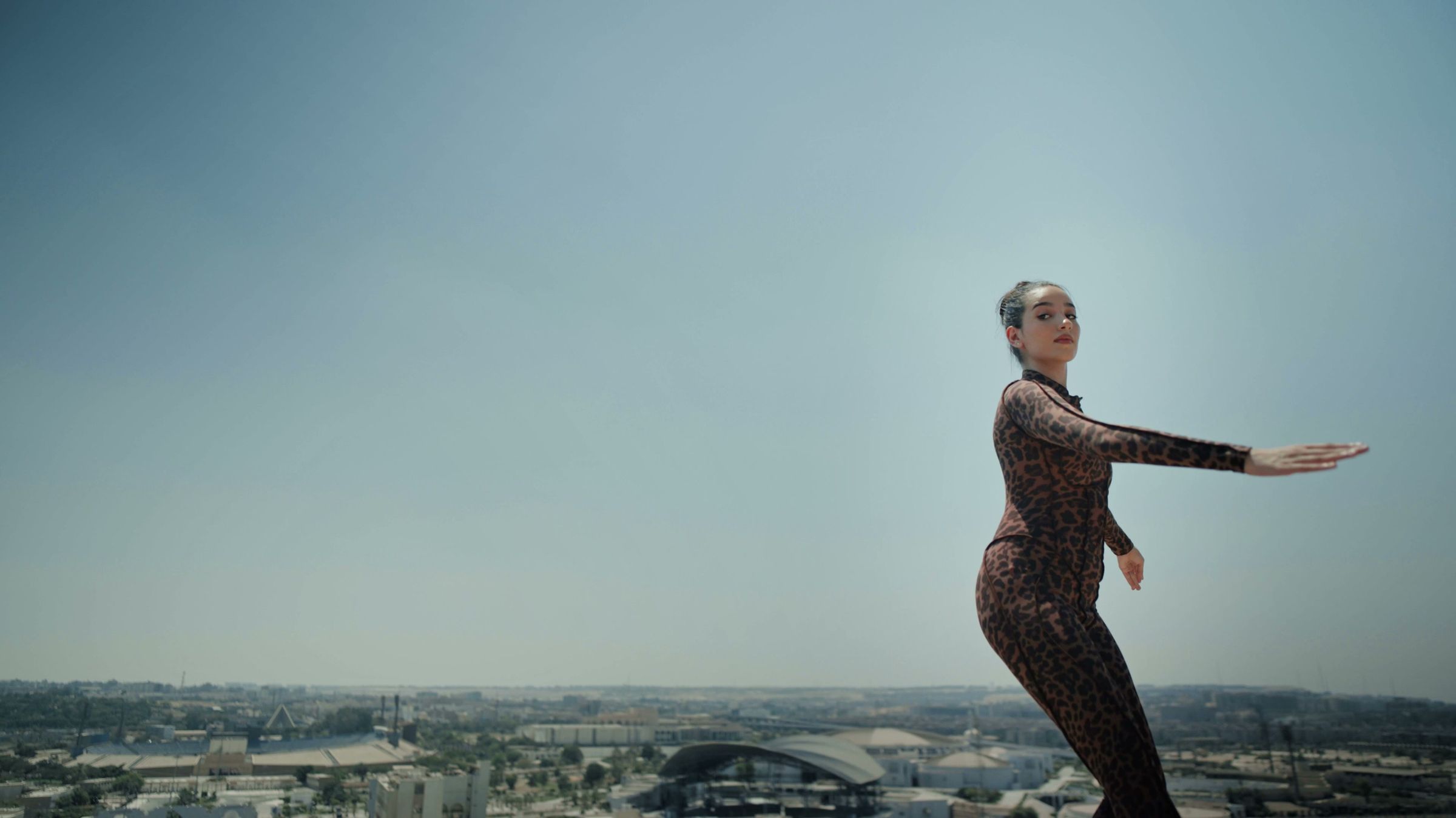 A woman in a leopard suit stretching on a rooftop above a wide, calm horizon